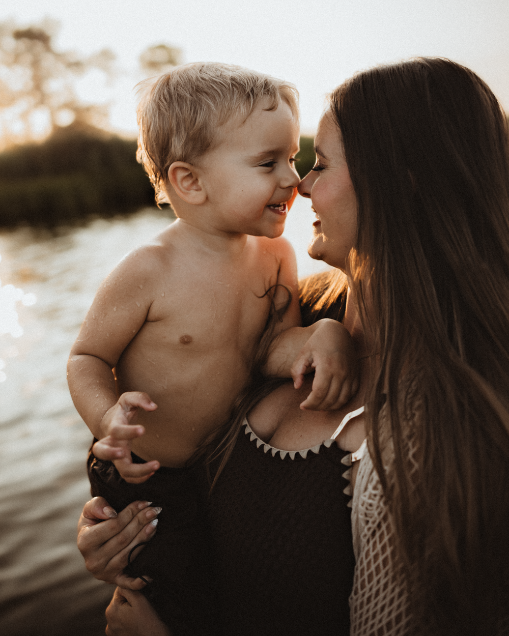 A mother holds her laughing toddler boy near a sunlit lake, sharing a tender nose-to-nose moment at golden hour.