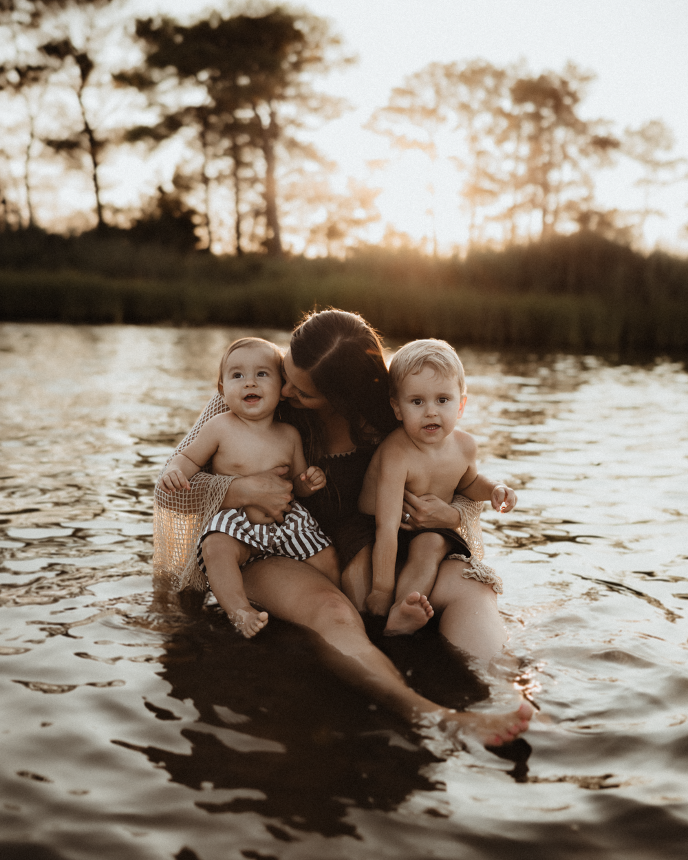 Mother sitting in a calm lake at golden hour, kissing one of her two toddlers as sunlight filters through trees.