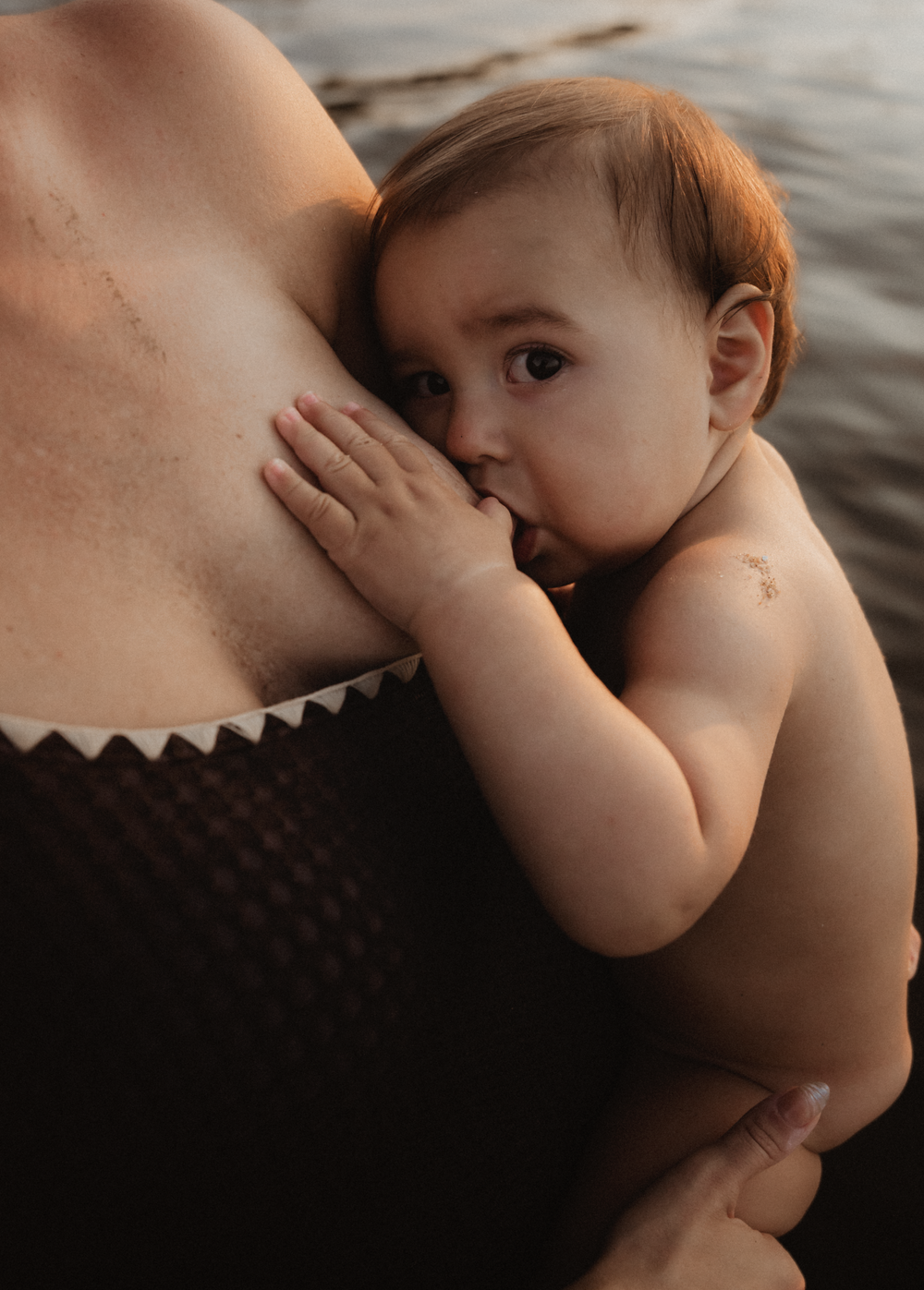 A baby breastfeeding while being held by its mother outdoors near water.