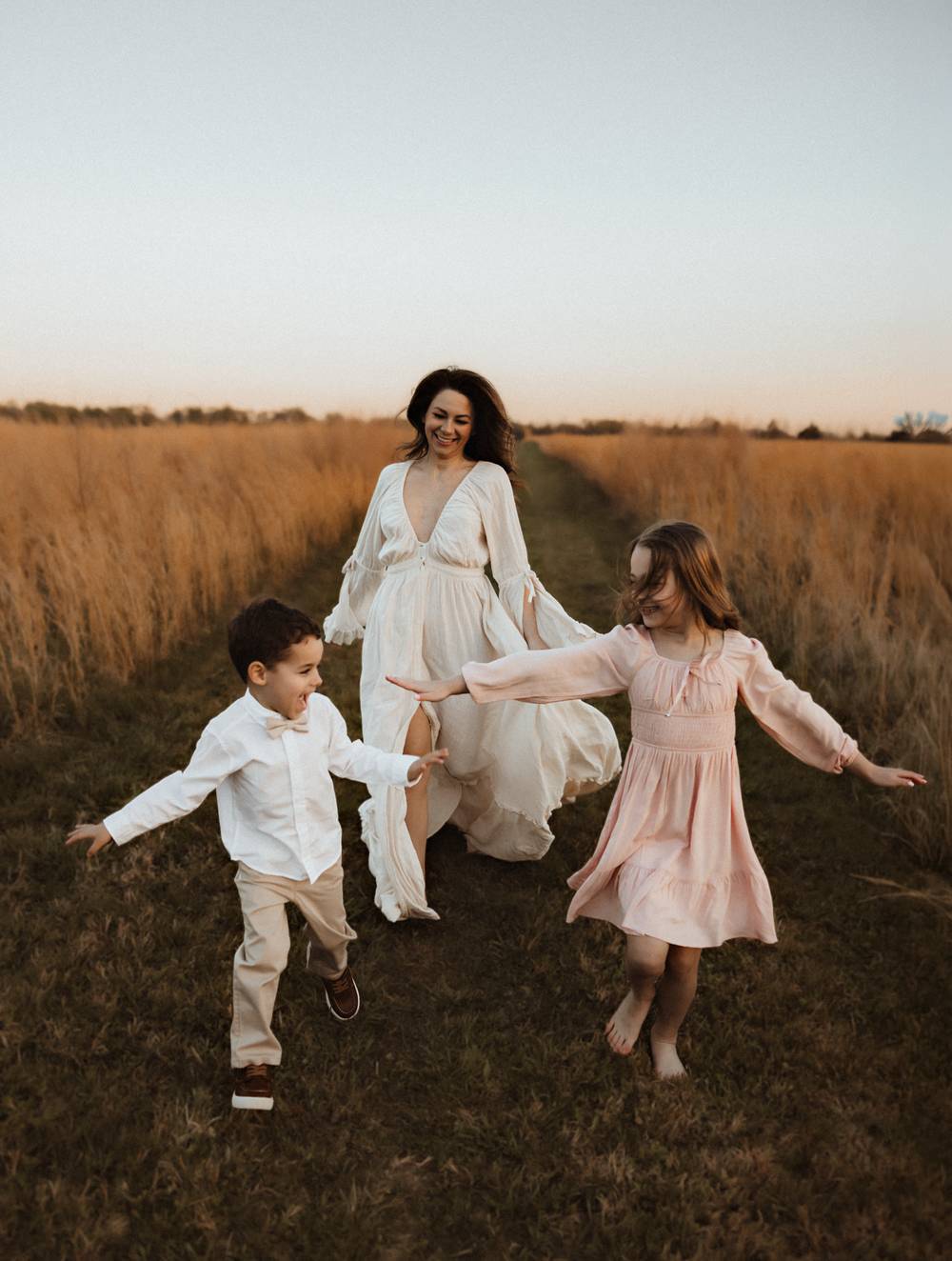 A woman in a white dress walks with two children through a golden field at sunset, holding hands and smiling.
