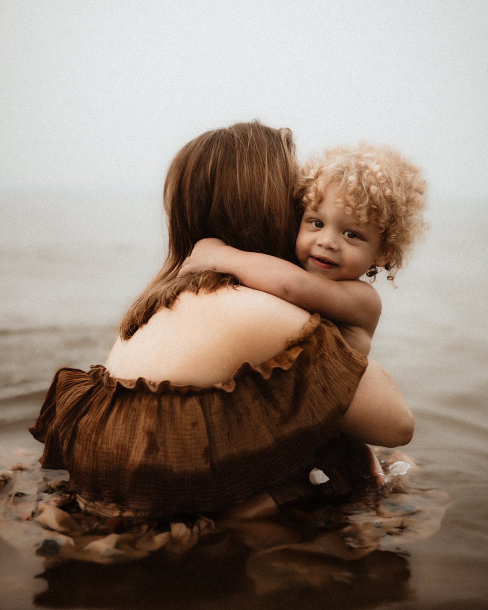 A mother and curly-haired toddler share a warm embrace while sitting in shallow ocean water.