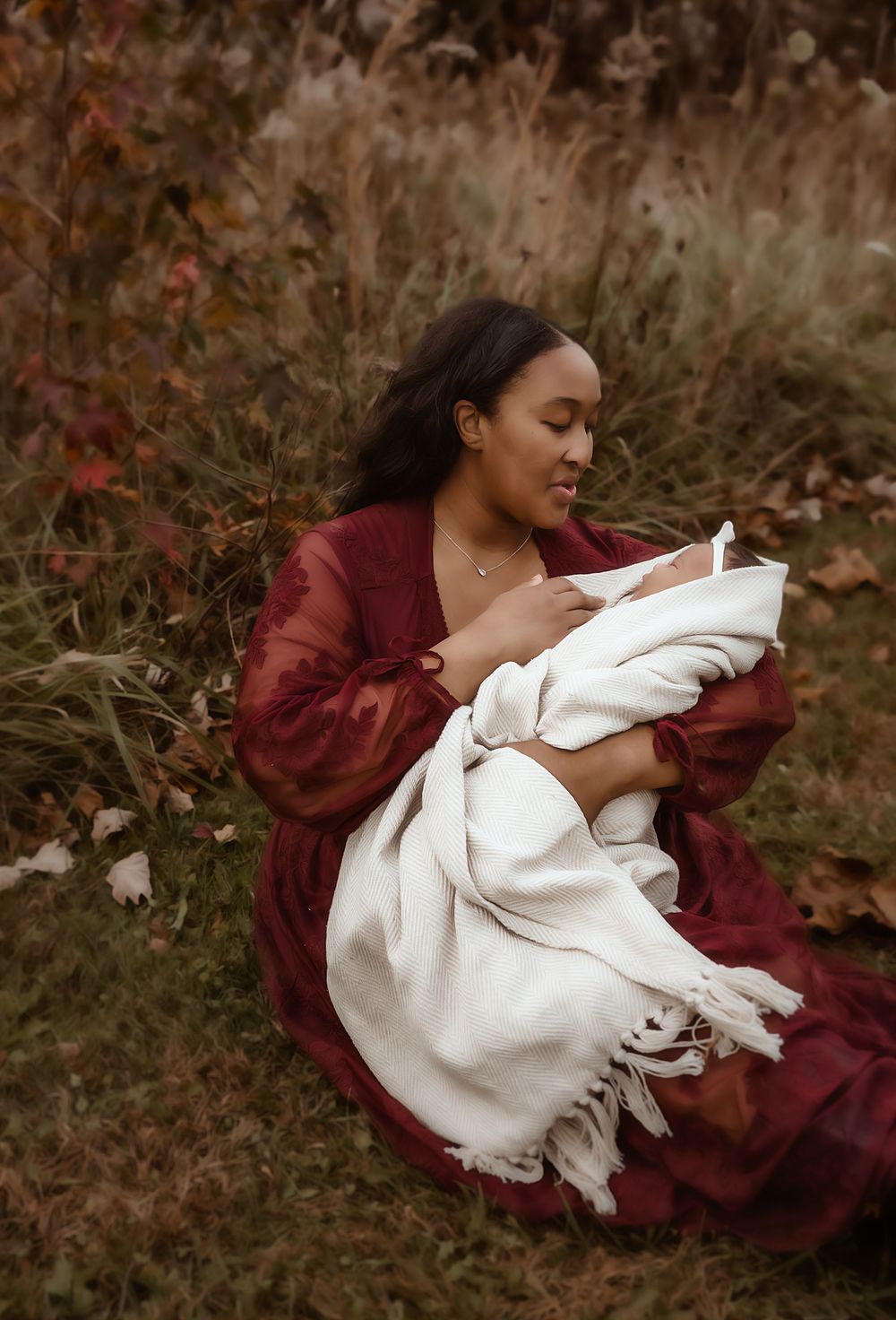 A mother in a red dress sits in an autumn field, tenderly cradling her newborn wrapped in a white blanket.