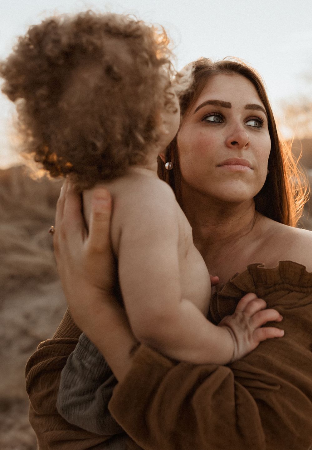 A mother tenderly holds her curly-haired toddler outdoors at golden hour, wearing a brown dress.