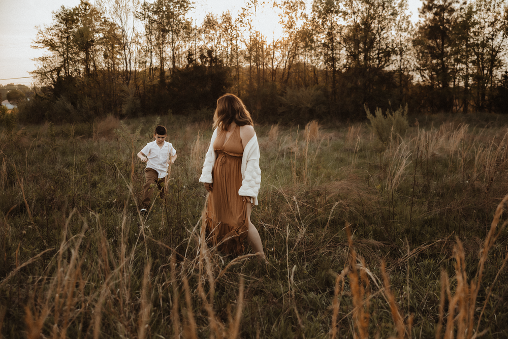A couple stands in a golden field at sunset, woman in brown dress and white jacket, man in white shirt behind her.