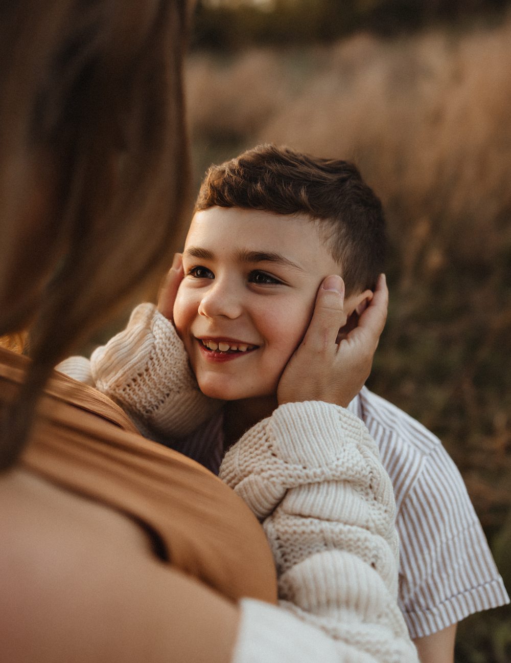 A smiling boy has his cheeks gently cupped by a woman's hands outdoors in a golden field.