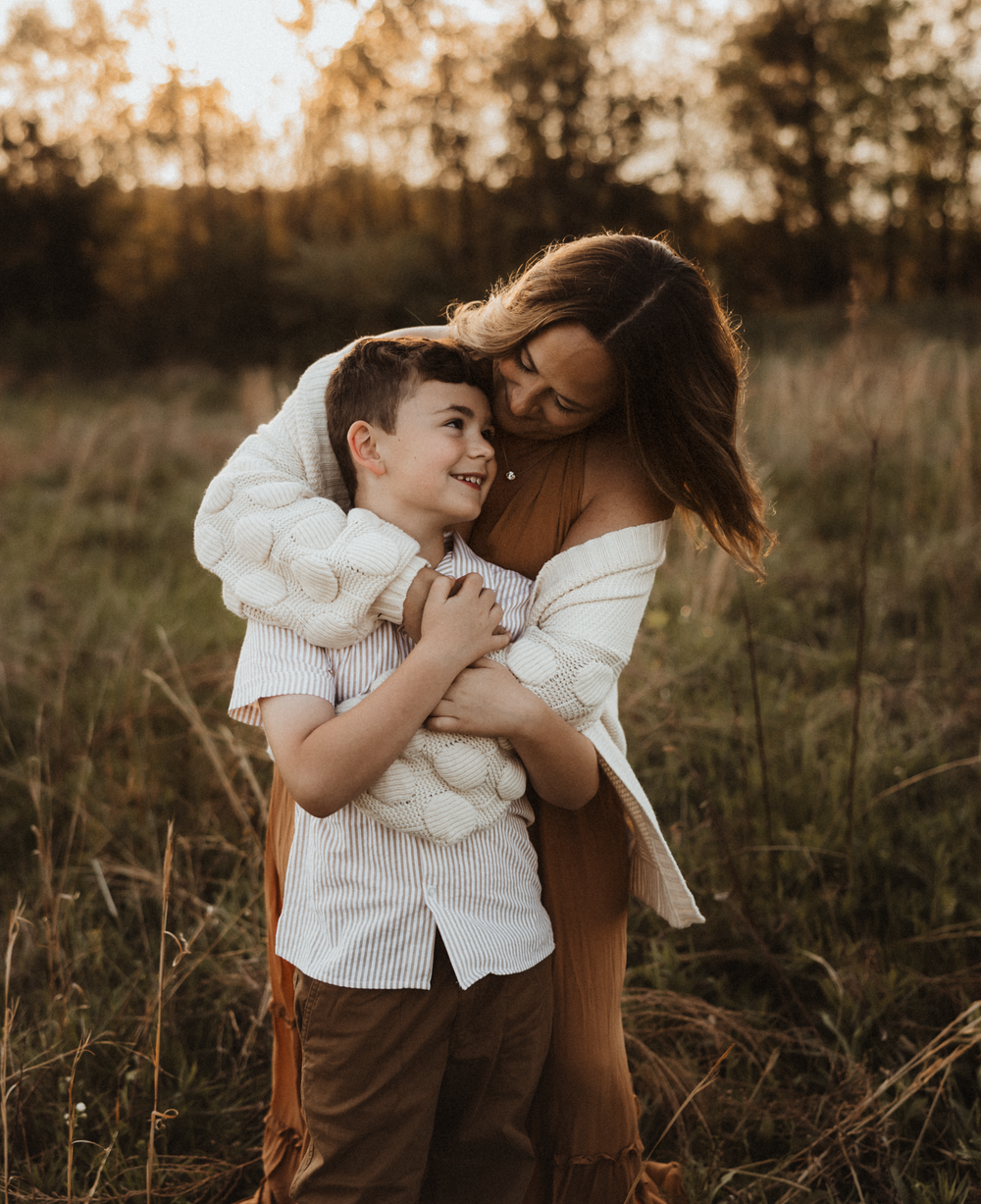 A mother tenderly embraces her smiling young son in a golden sunlit field, both wearing warm earth-toned outfits.
