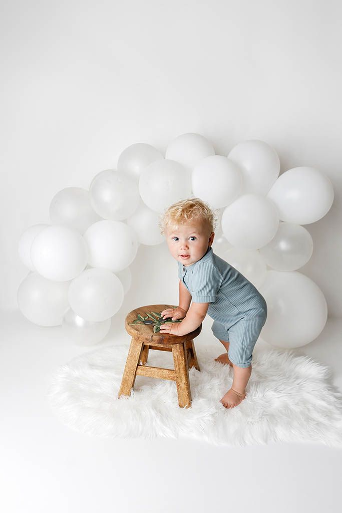 Toddler in blue romper standing by wooden stool surrounded by white balloon arch on fluffy white rug.