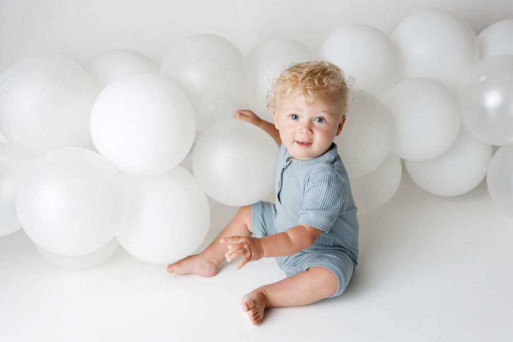 Smiling toddler with curly blonde hair in a blue romper sitting among white balloons on a white background.