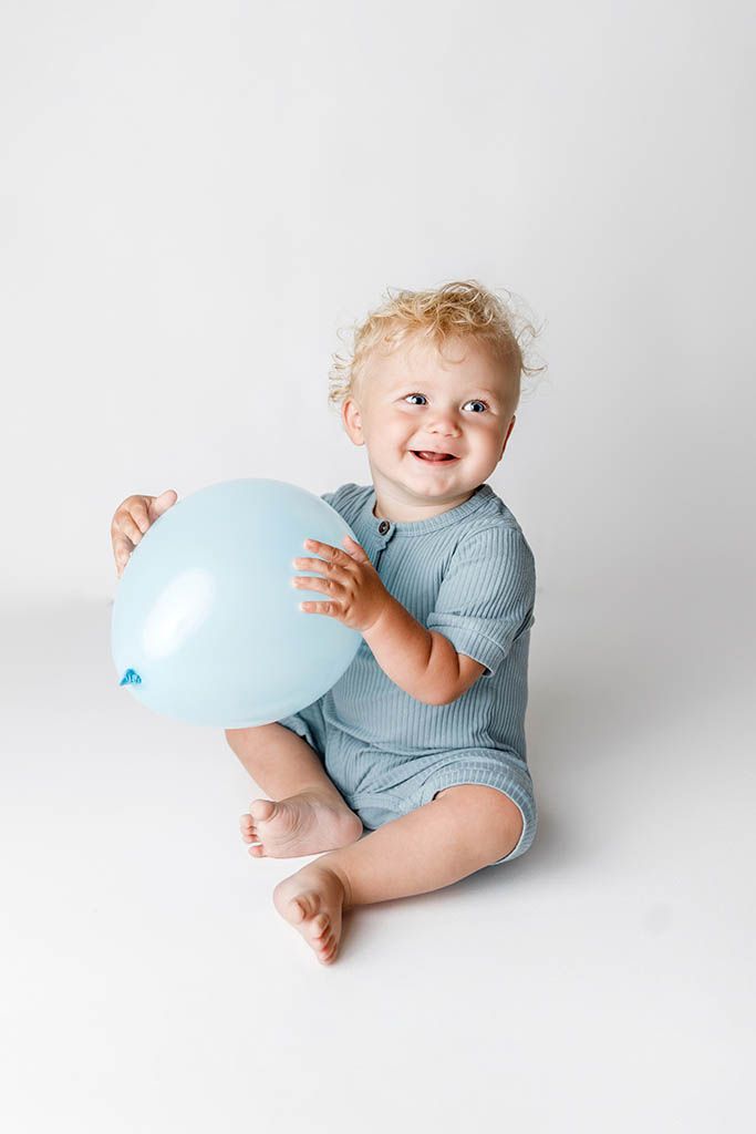 Smiling baby with curly blonde hair sitting on white background holding a light blue balloon, wearing a blue romper.
