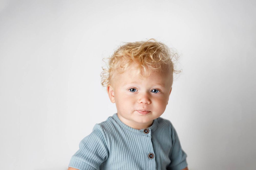 Cute toddler with curly blonde hair and blue eyes wearing a sage green ribbed outfit against a white background.