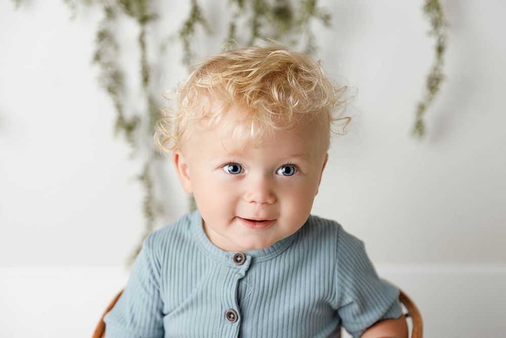 Smiling toddler with curly blonde hair wearing a sage green ribbed button-up shirt, posed against a white background.