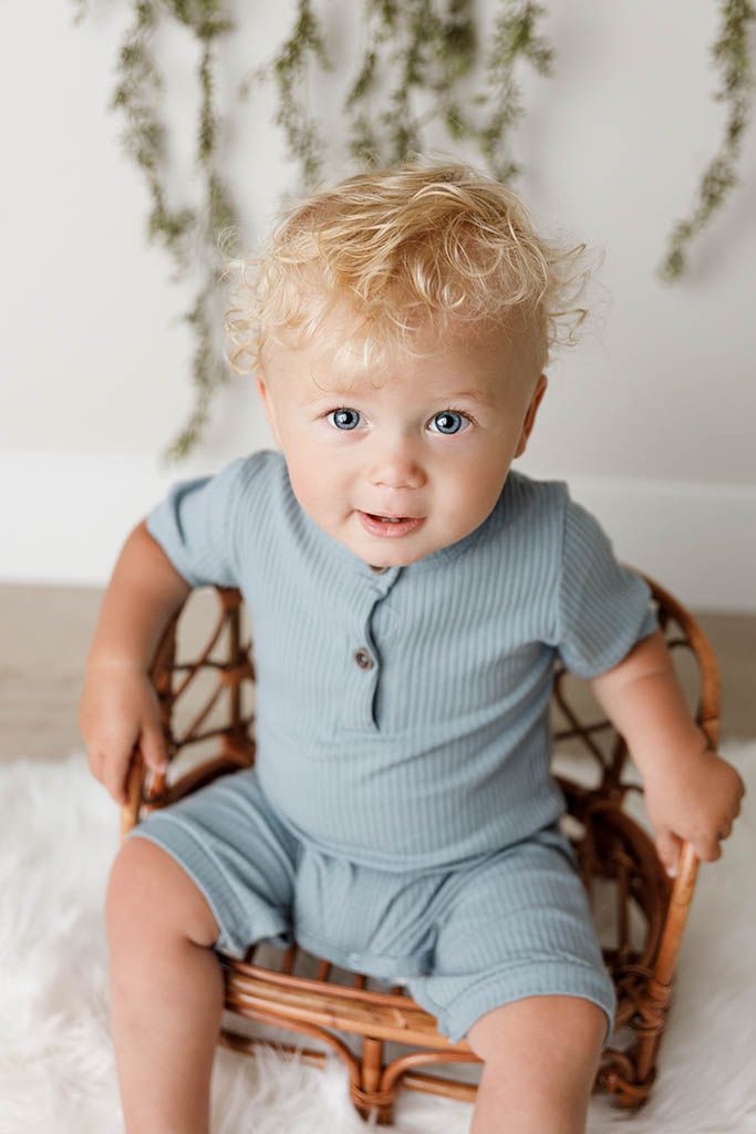 Adorable blonde curly-haired baby with blue eyes sitting in a rattan chair wearing a blue ribbed romper.