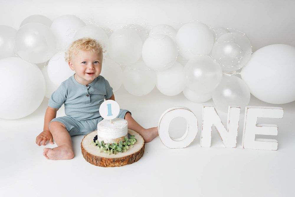 Smiling baby boy in blue romper sits beside a white cake and ONE sign, surrounded by white balloons for his first birthday.