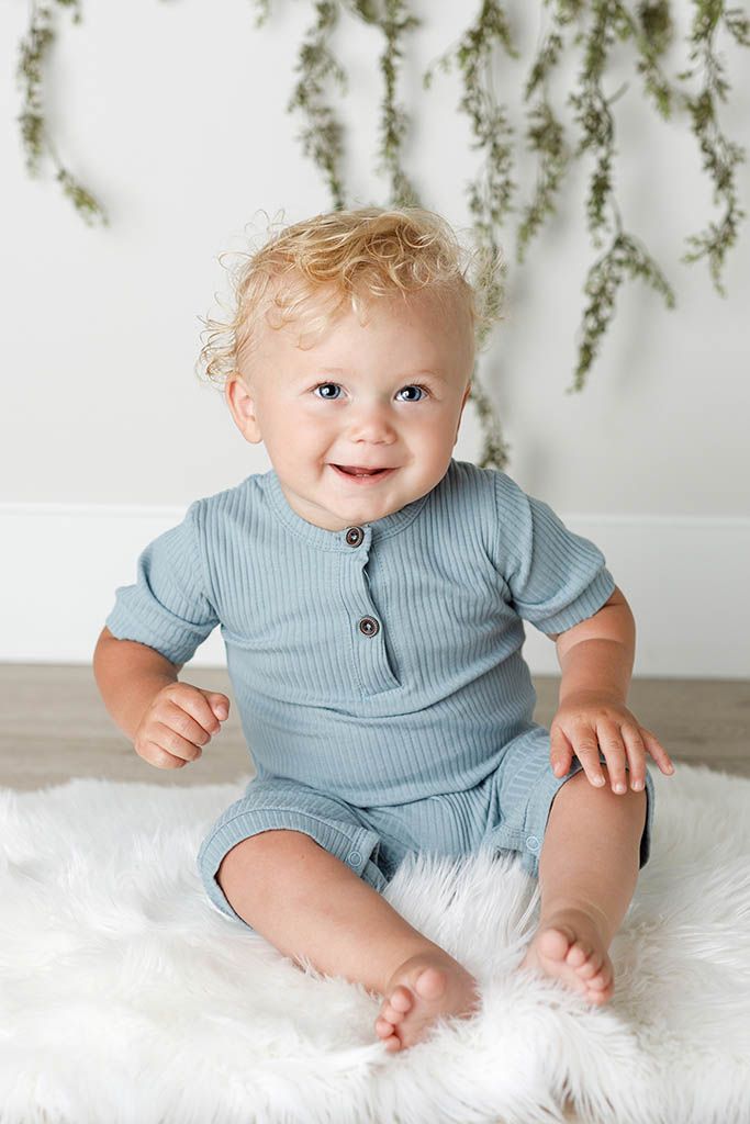 Smiling baby with curly blonde hair sits on white fur rug wearing a blue ribbed romper in a bright studio setting.