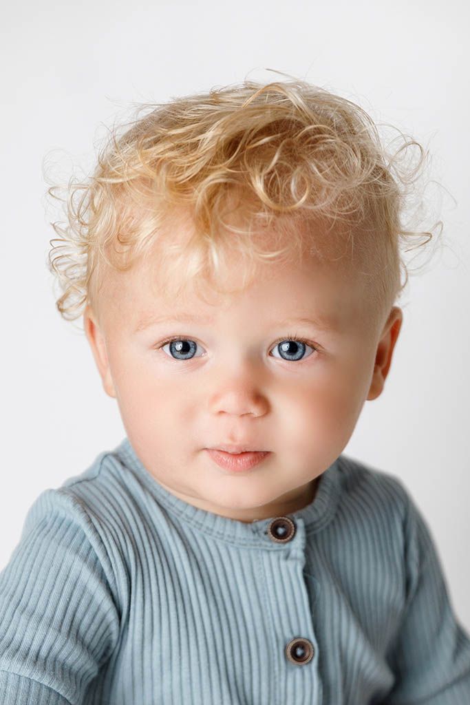 Close-up portrait of a cute toddler with curly blonde hair and blue eyes wearing a light blue ribbed top.