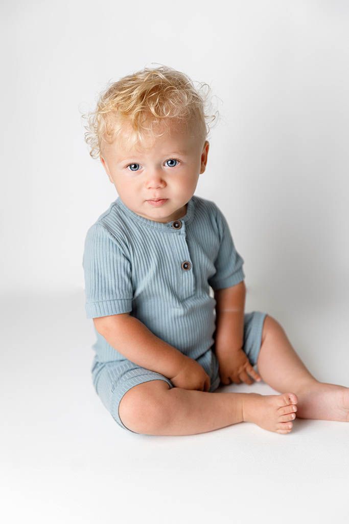 Cute toddler with curly blonde hair wearing a light blue ribbed romper sitting on a white background.