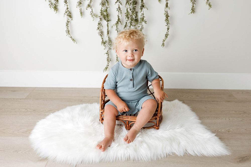 Adorable toddler with curly blonde hair sitting in a wicker chair on a white fluffy rug in a bright studio.