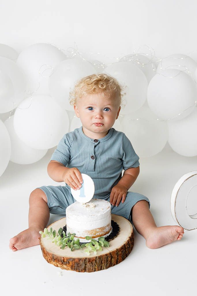 Toddler boy in blue romper smashing first birthday cake on wood slice surrounded by white balloons.
