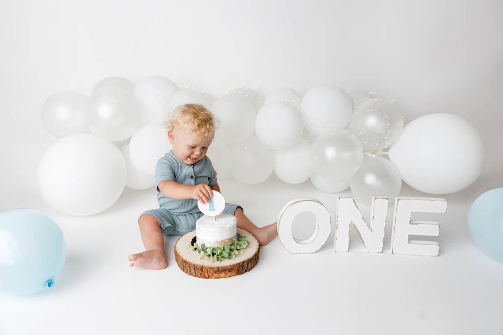 Toddler boy in blue romper touching first birthday cake surrounded by white and blue balloons with ONE letters.