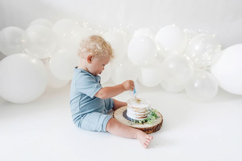 Toddler boy in blue romper smashing first birthday cake surrounded by white balloons on white background.