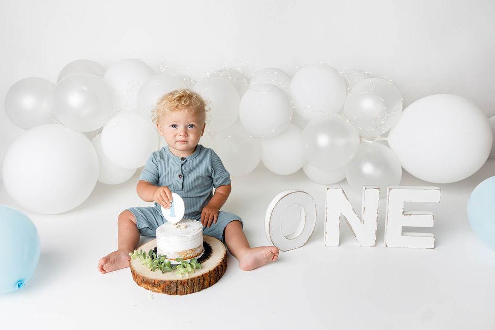 Toddler boy sitting beside a white cake on a wood slice for a first birthday cake smash with white balloons.