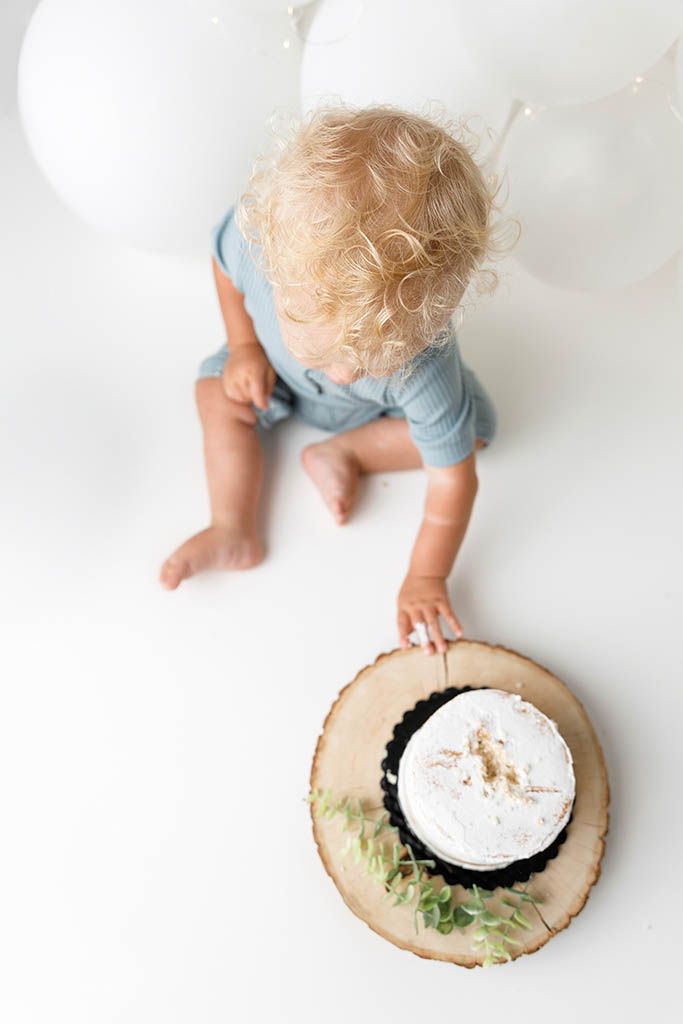 Toddler with curly blonde hair in blue romper reaching for a white smash cake on wood slice with balloons.