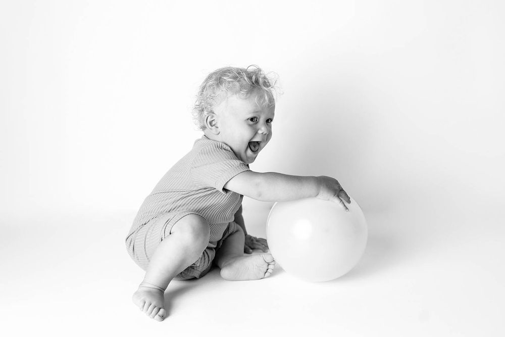 Black and white photo of a cute toddler sitting on floor playing with a white ball.
