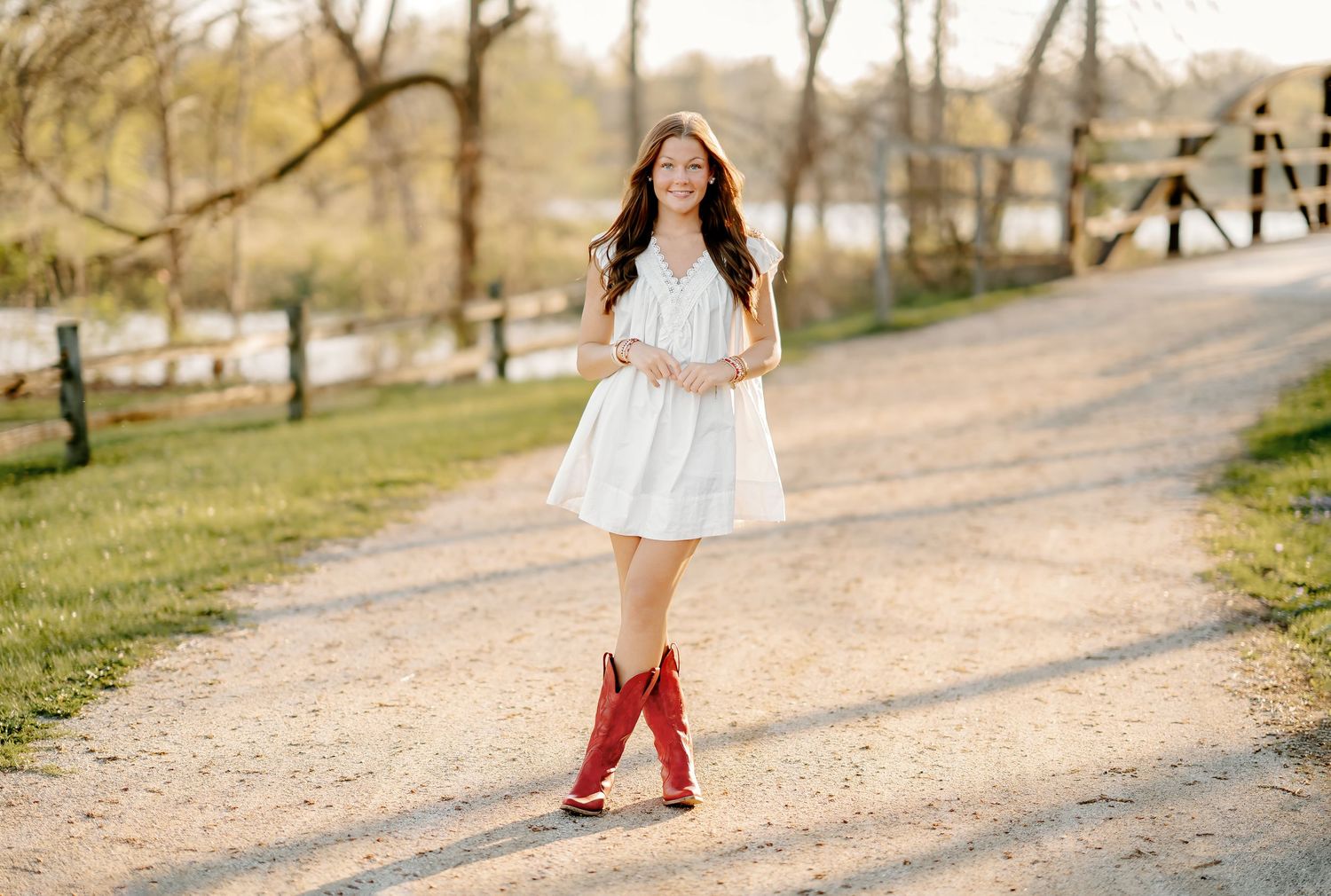 Young woman in white dress and red cowboy boots smiling on a scenic gravel path surrounded by trees.