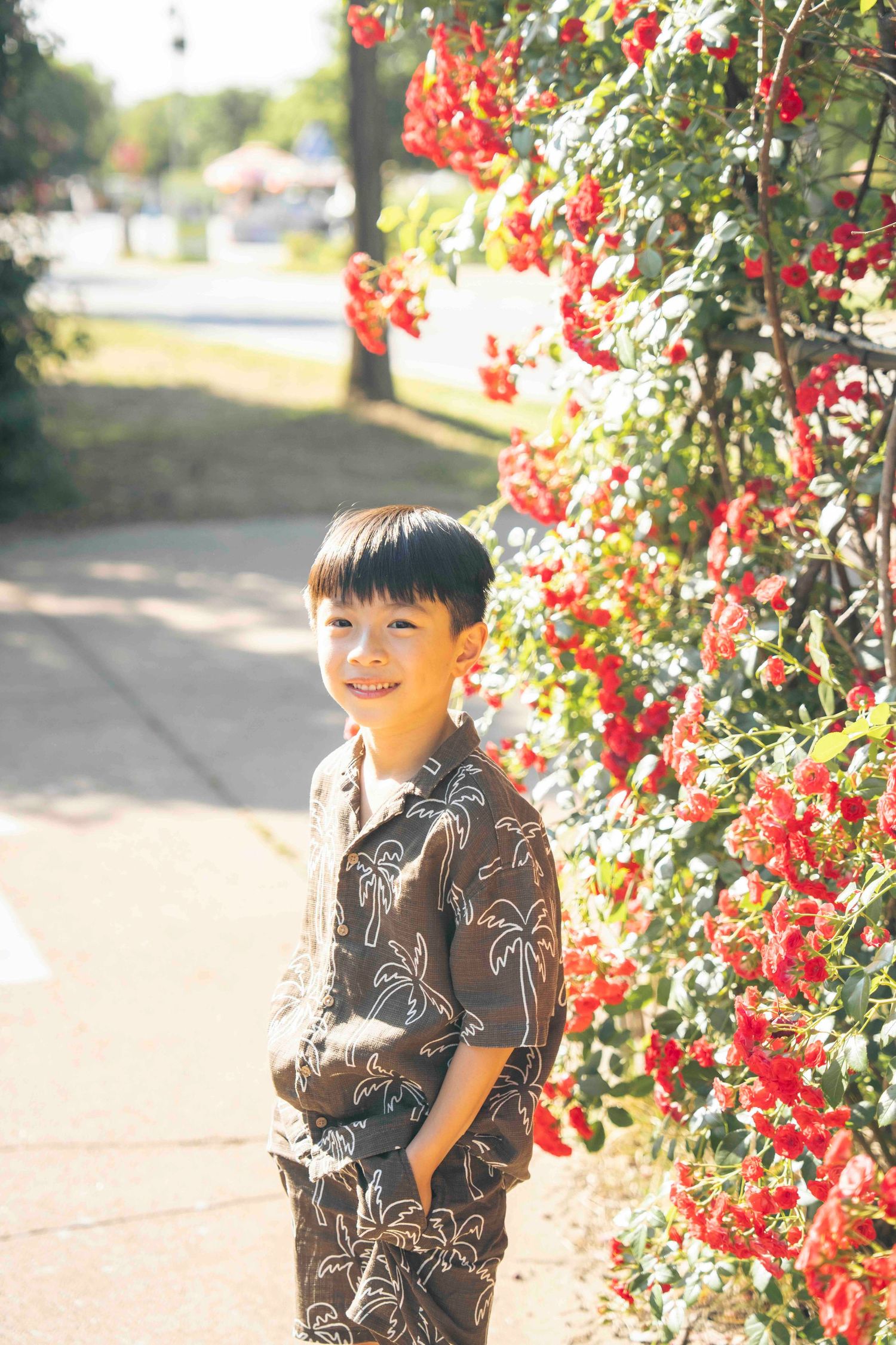 Smiling Asian boy in a brown Hawaiian shirt standing beside vibrant red flowering bushes on a sunny day.