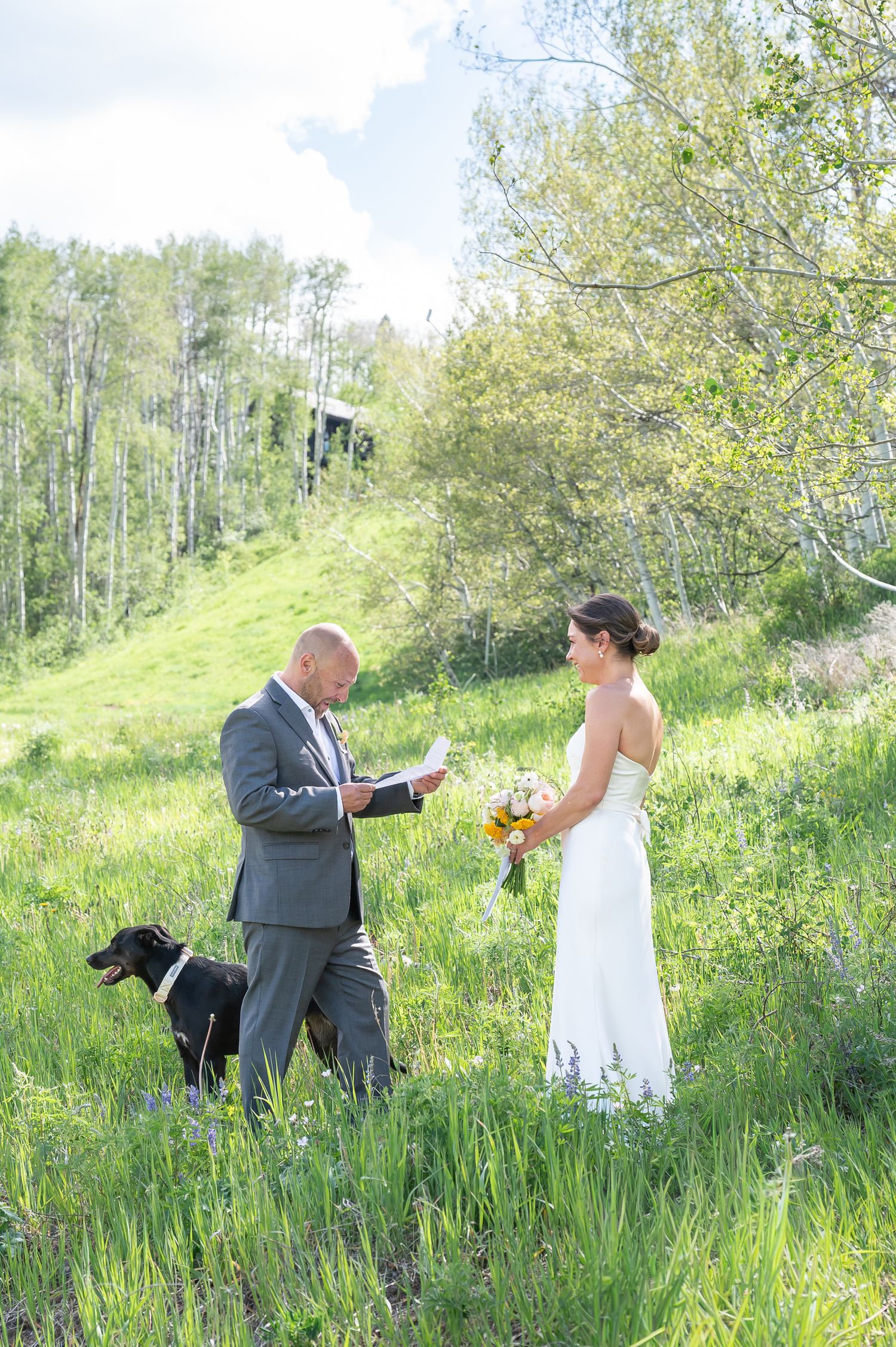 Outdoor Colorado wedding ceremony in a lush green meadow with groom reading vows, bride holding bouquet, and black dog nearby.