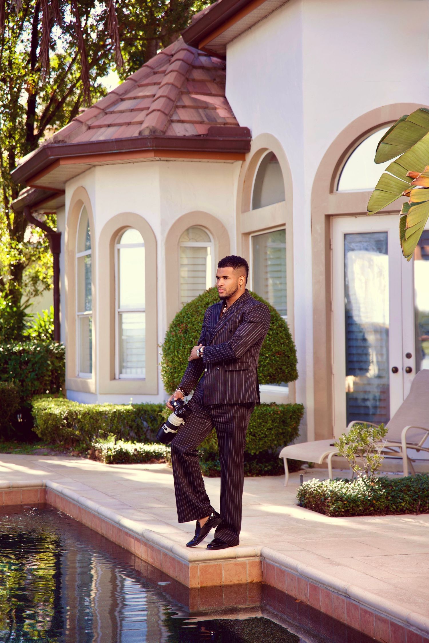 Man in pinstripe suit holding camera stands by pool outside elegant estate with arched windows and tiled roof.