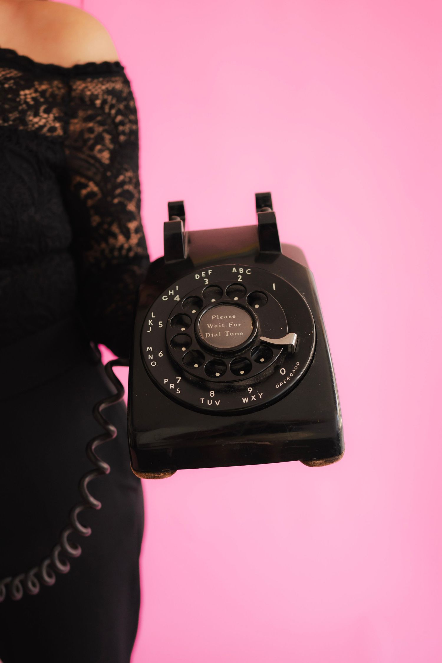 Woman in black lace dress holding a vintage black rotary dial telephone against a pink background.