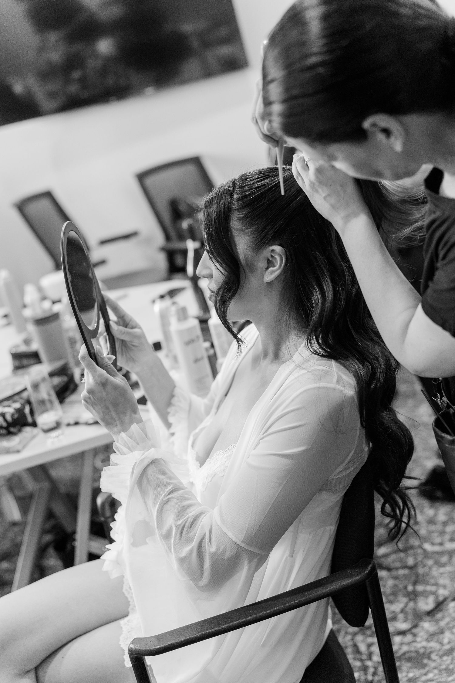 Black and white photo of a bride holding a mirror while a stylist arranges her long wavy dark hair.