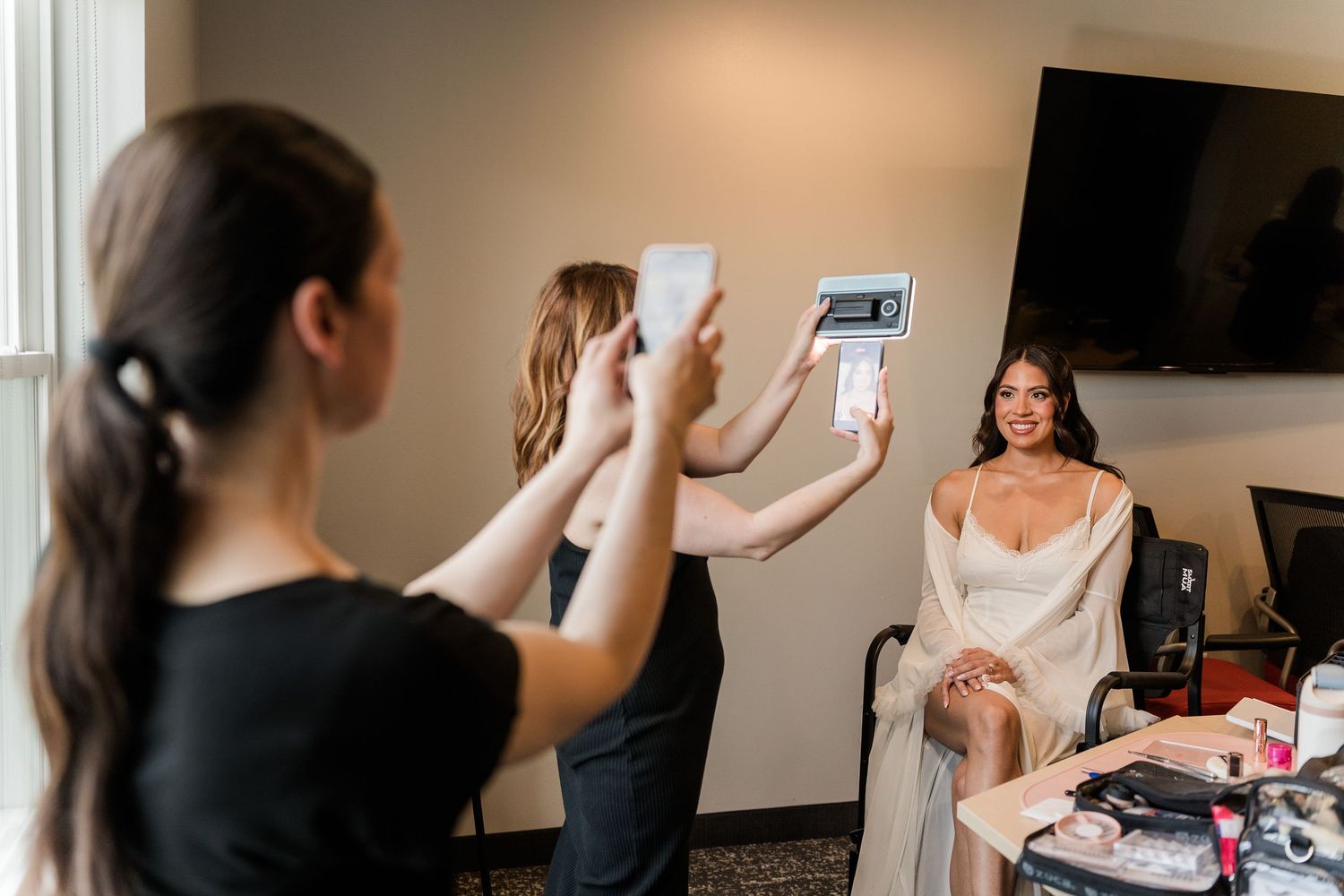 Bridesmaids photographing a smiling bride in a white robe during wedding morning preparations indoors.
