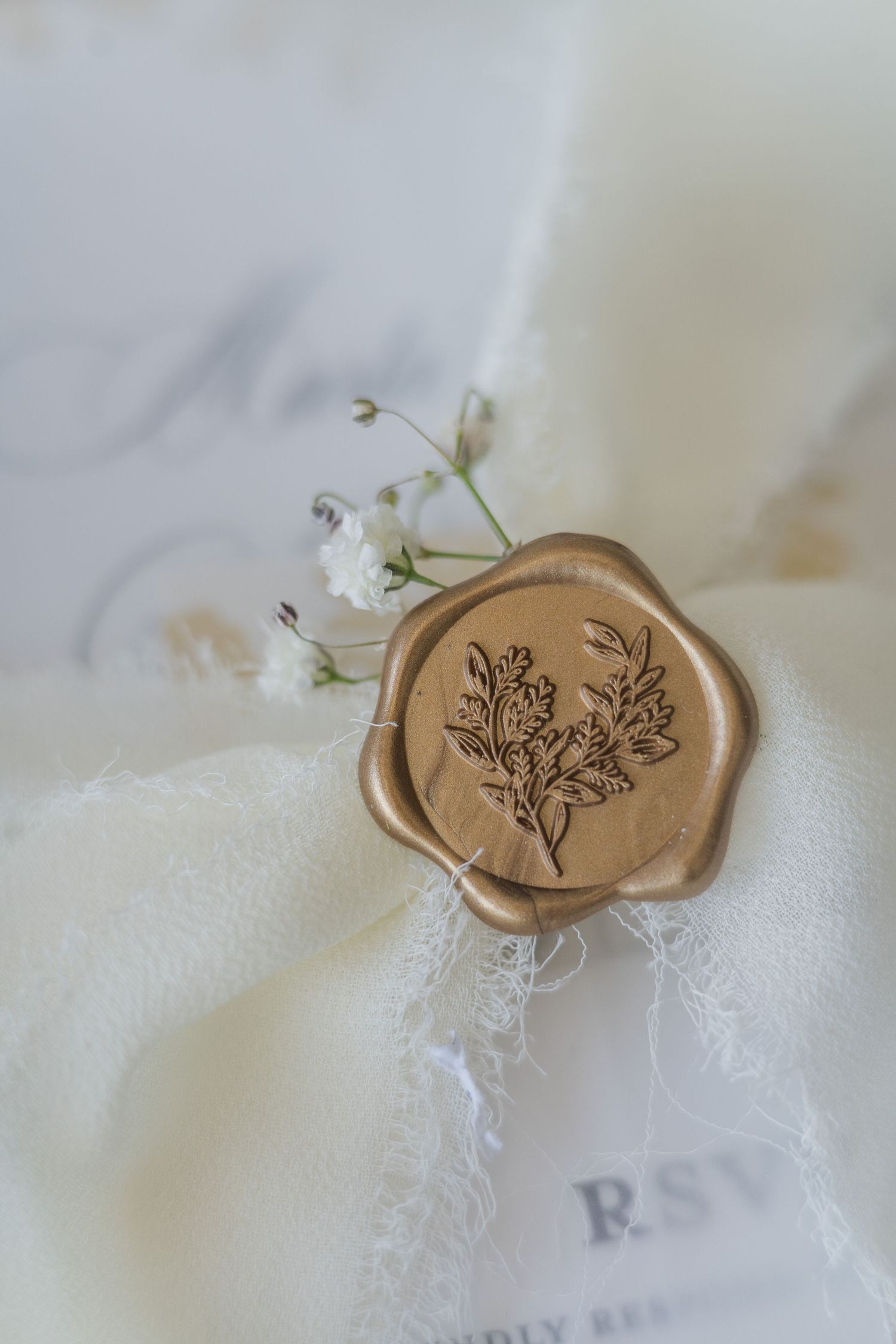 Gold floral wax seal on ivory fabric beside white baby's breath flowers and RSVP card.