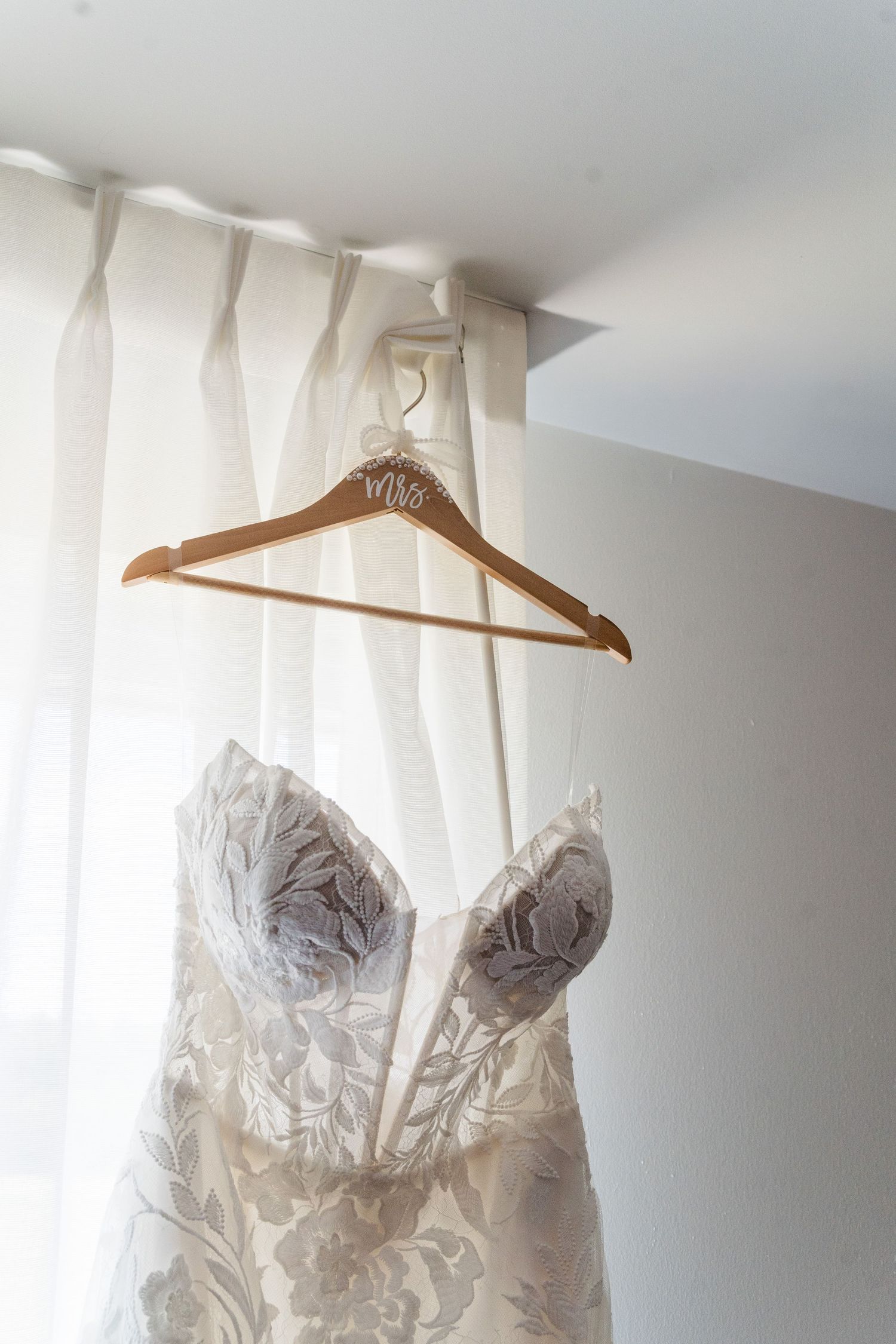 Elegant lace wedding dress hanging on a wooden Mrs. hanger against a bright white curtained window.