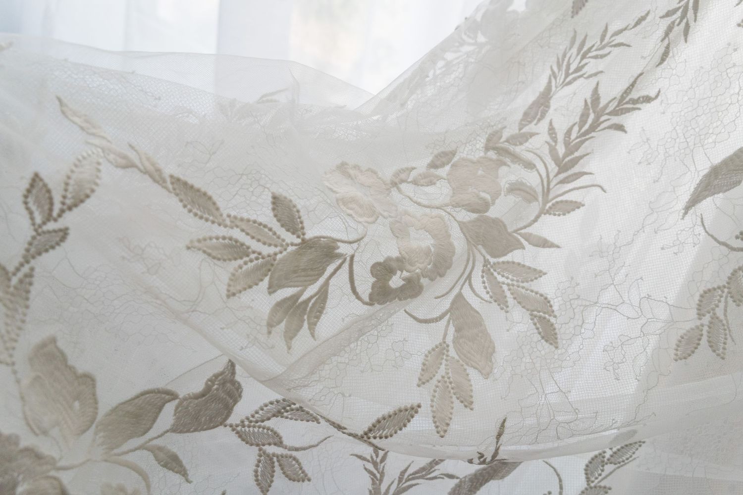 Delicate sheer white fabric with intricate embroidered floral and leaf pattern in soft taupe tones for bridal decor.