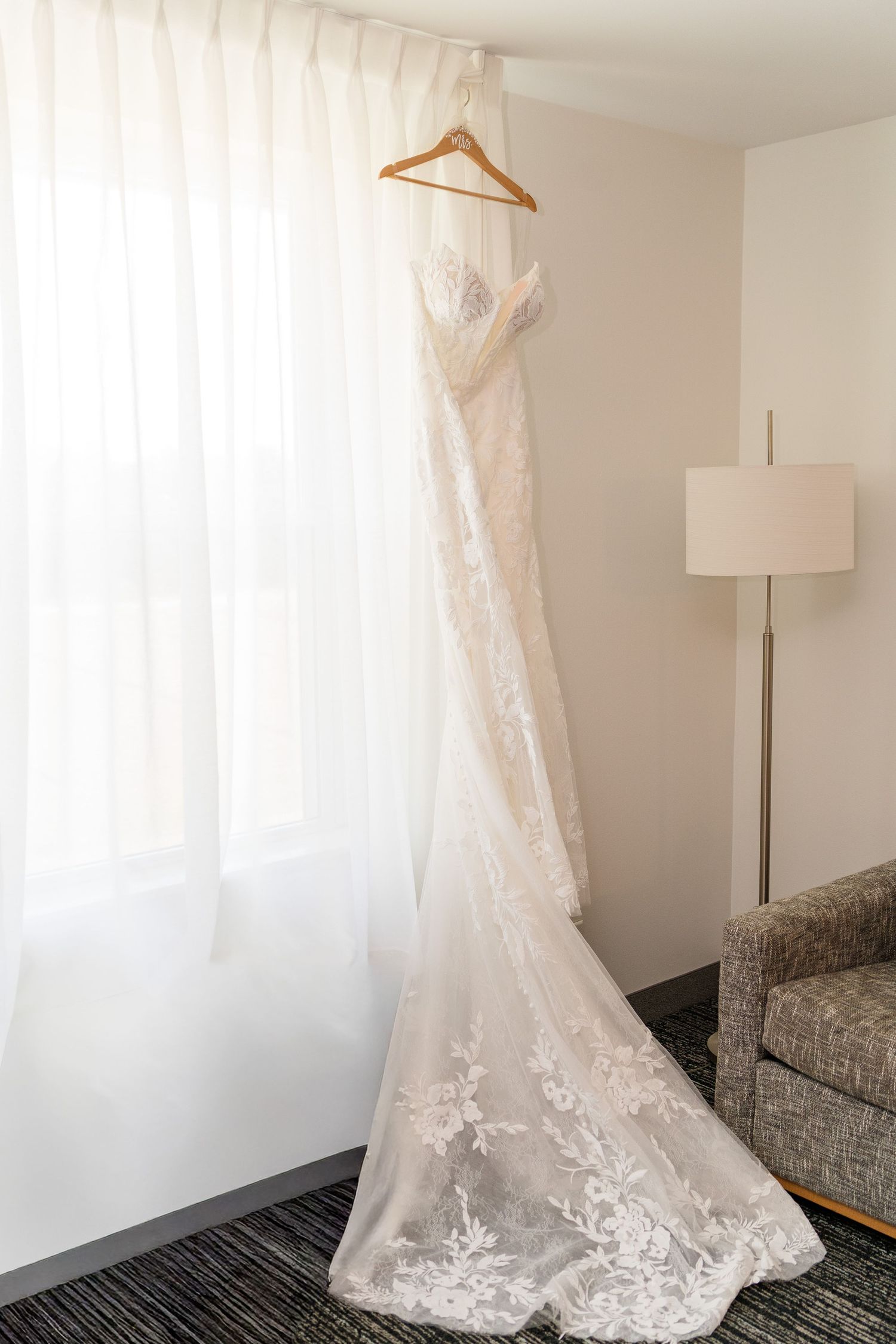 Elegant lace wedding dress hanging on a wooden hanger in front of sheer white curtains in a hotel room.