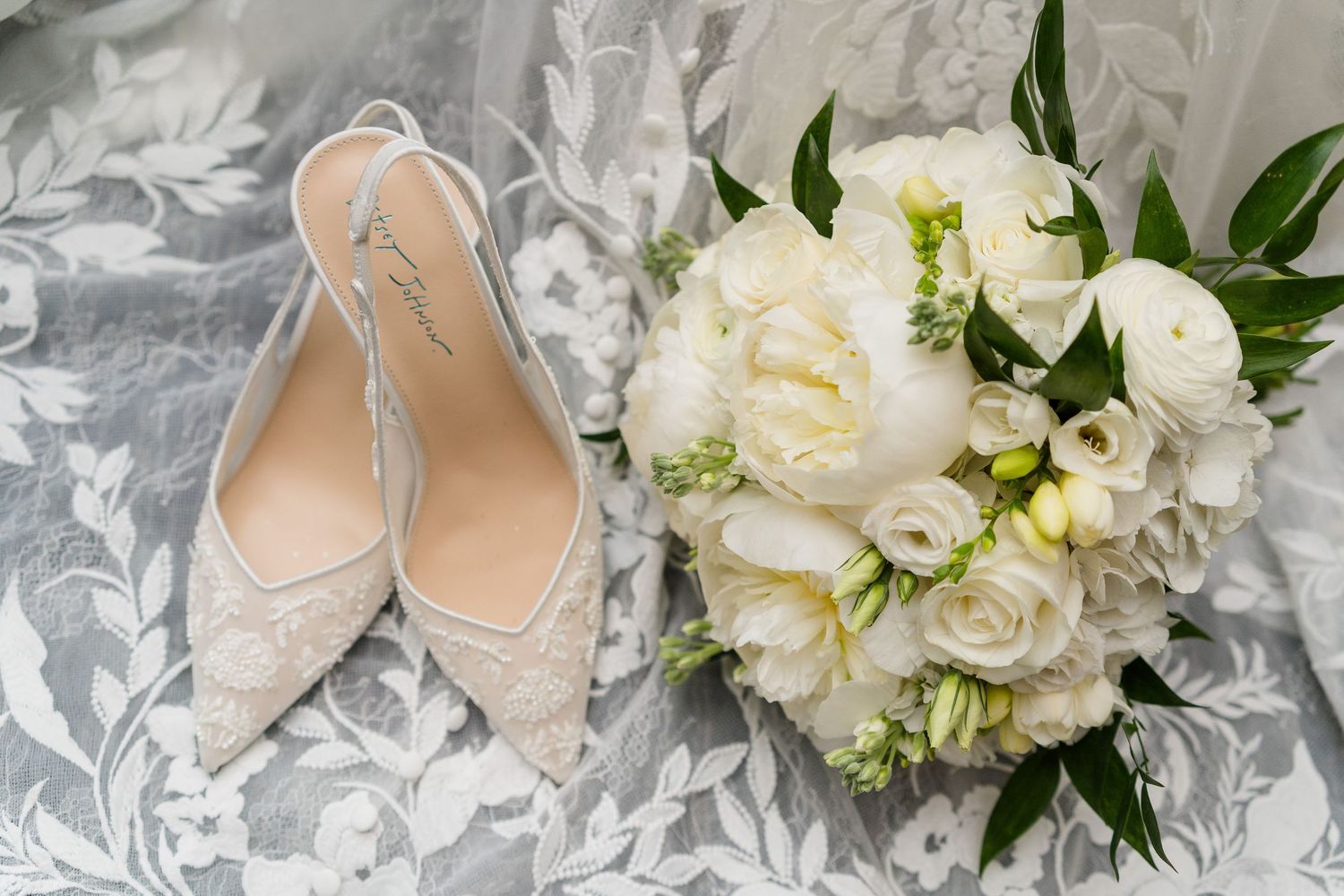 White lace bridal heels beside a lush bouquet of white peonies and ranunculus on a gray lace fabric.