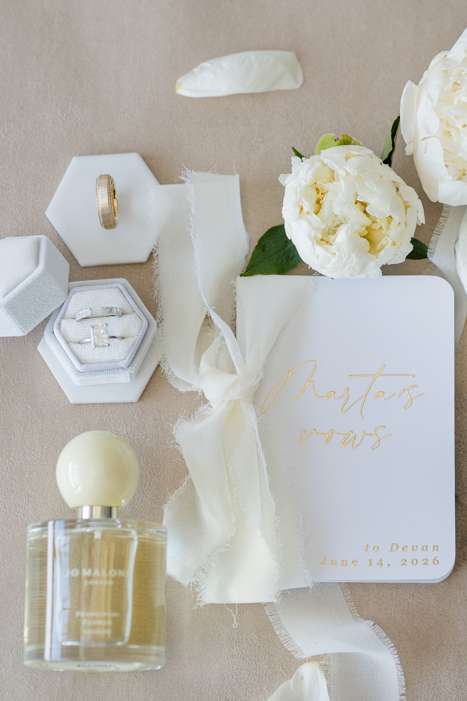Elegant wedding flat lay with vow booklet, diamond ring, gold band, Jo Malone perfume, white peonies, and ribbon on beige backdrop.