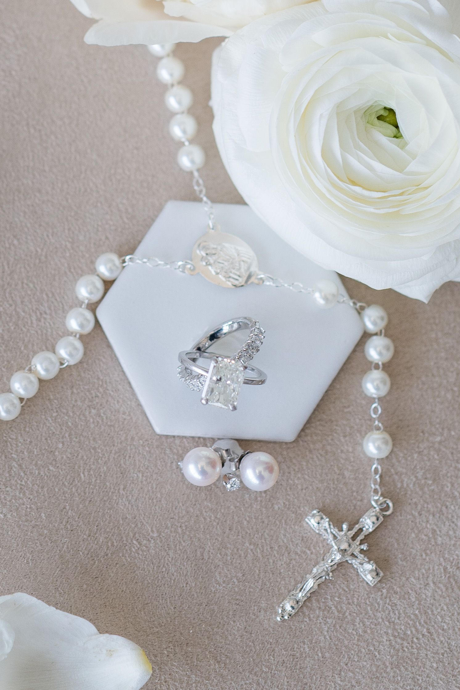 Pearl rosary with silver crucifix, diamond ring, pearl earrings on white hexagon tile with white roses.