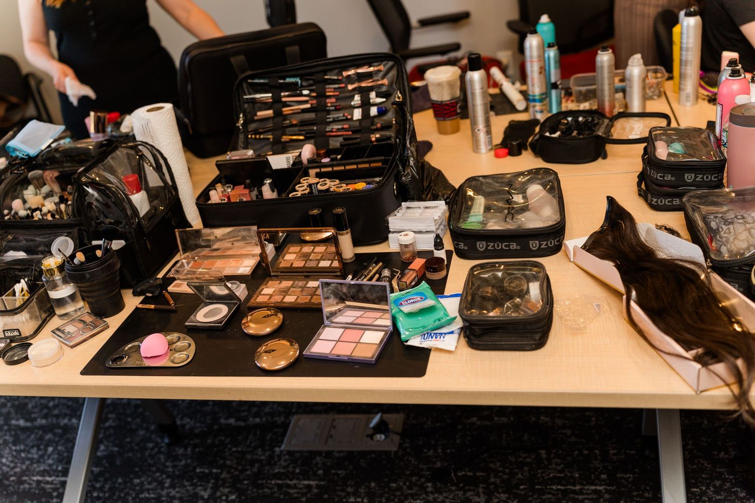 A professional makeup artist table covered with open cosmetic cases, palettes, brushes, and beauty products backstage.