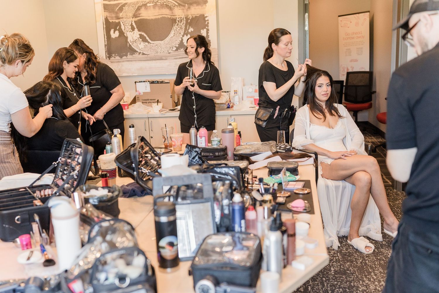 Bridesmaids getting hair and makeup done in a busy bridal suite with beauty products on a table.