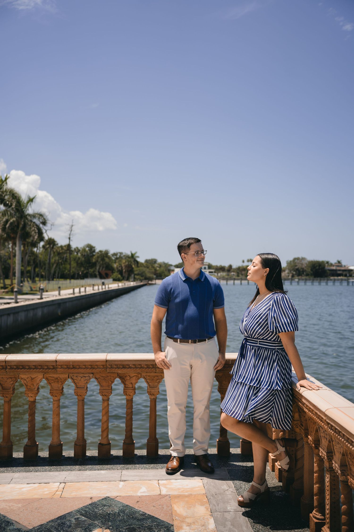 Couple posing on ornate stone balcony overlooking waterway with palm trees under blue sky.