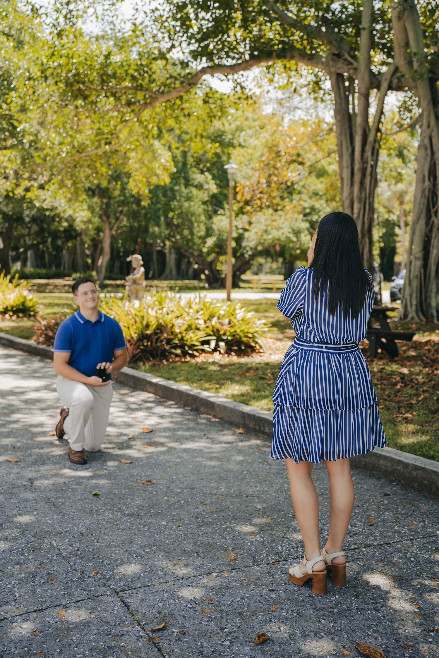 Man kneeling on one knee proposing to woman in striped dress in a sunny park setting.