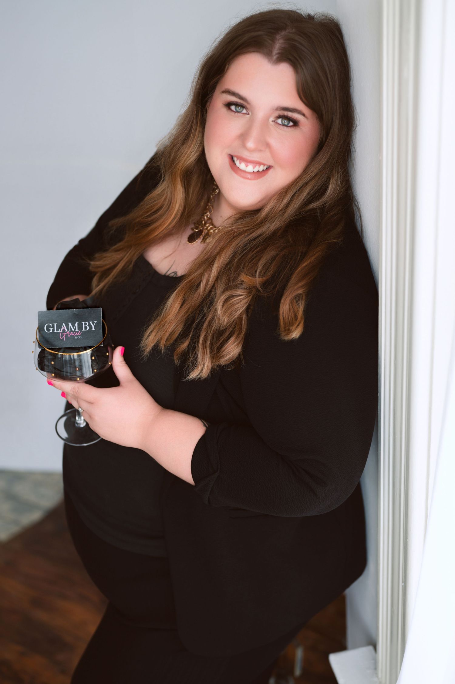 Smiling woman in black outfit holding a Glam By wine glass, posing elegantly against a white wall.