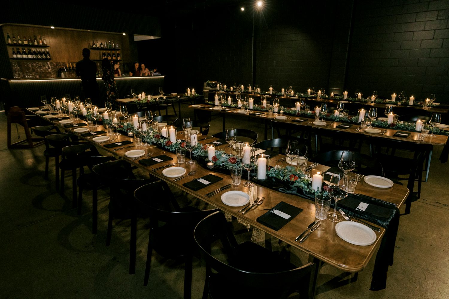 Elegant dark dining room with long wooden tables set with candles, greenery runners, and white plates for an intimate event.