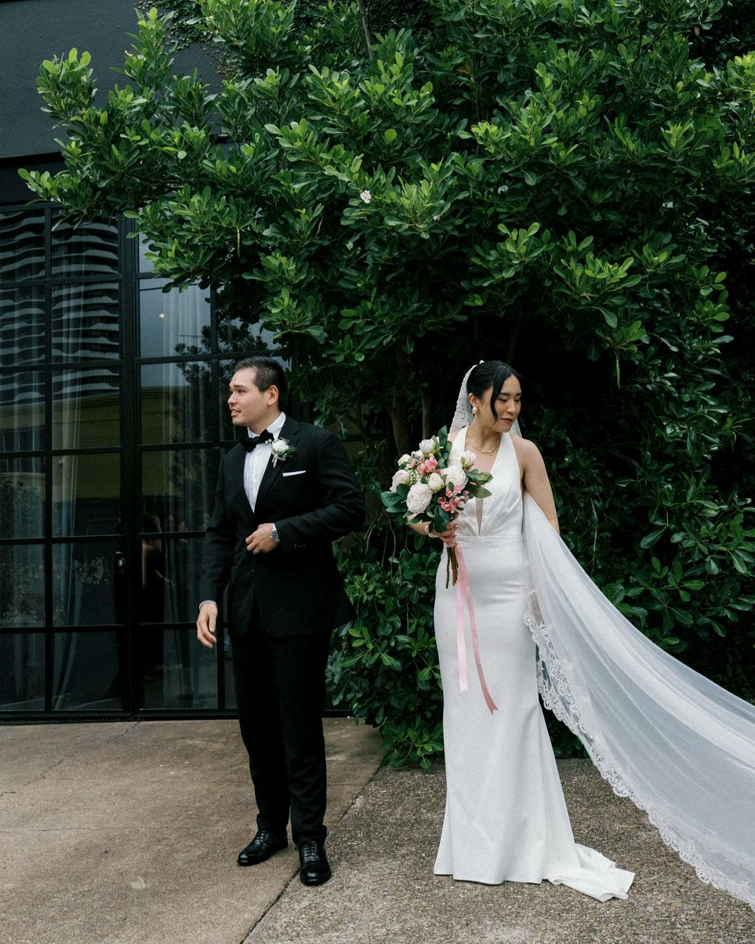 Bride in white gown with veil holds bouquet beside groom in black tuxedo, posing before lush green foliage. the bethnal. Newstead Brisbane