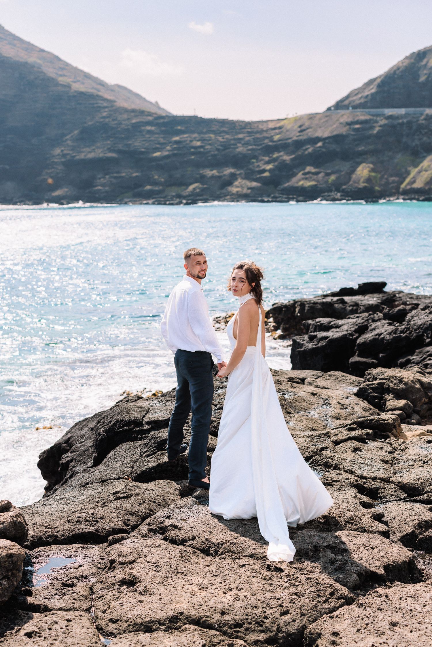 A bride and groom stand on rocky coastal cliffs with sparkling blue ocean and green mountains behind them.