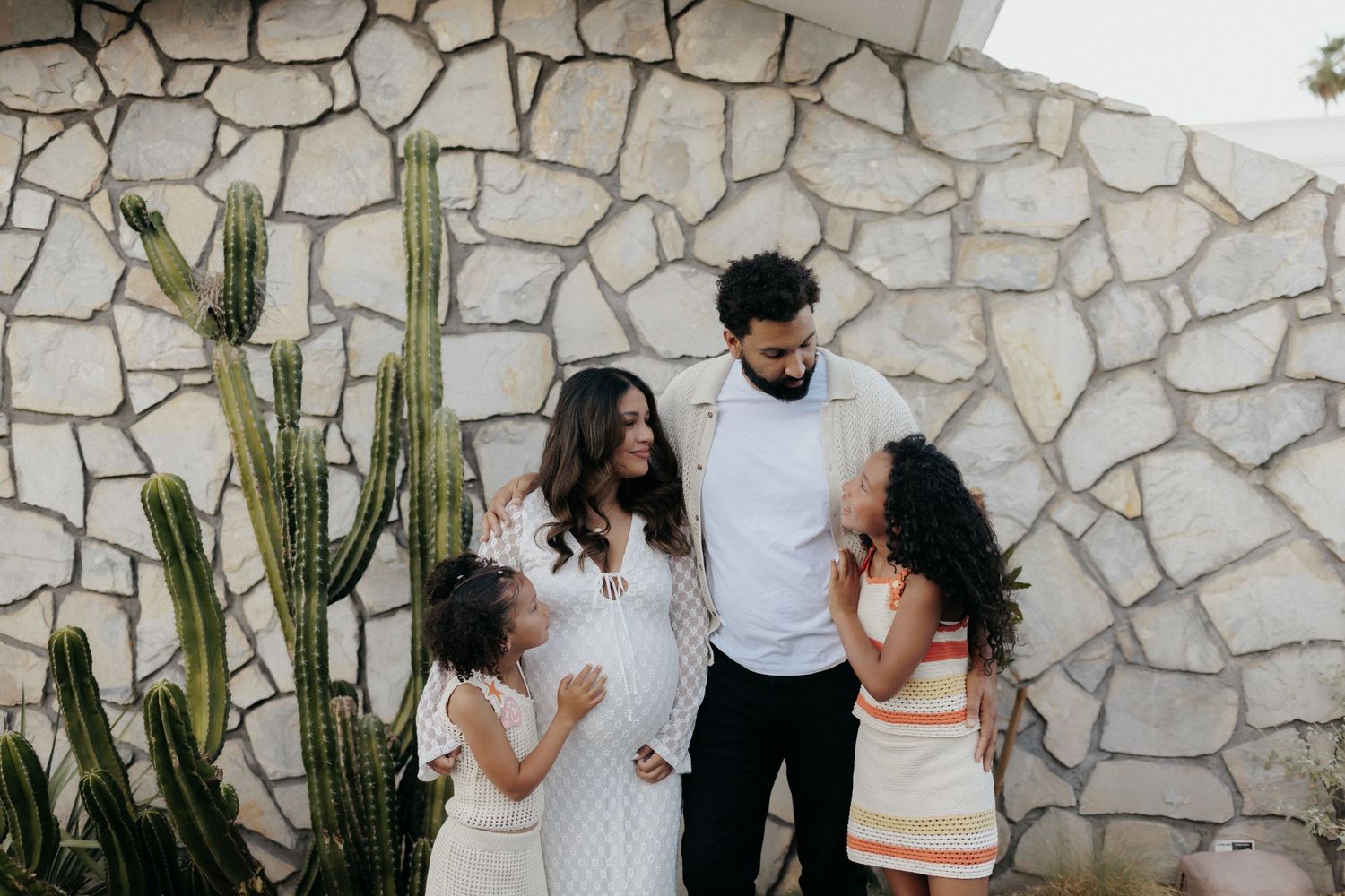 A family of four poses happily against a stone wall with a cactus, featuring a pregnant woman in a white dress.
